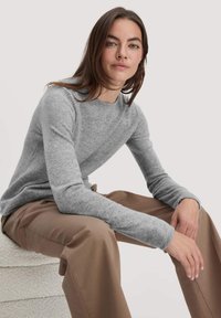 Grauer Strickpullover mit Rundhalsausschnitt und langen Ärmeln, kombiniert mit braunen Hosen. Weiche Textur und schlichtes, sauberes Design.