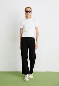 Young person with short blonde hair wearing black sunglasses, white t-shirt, black pants, and beige sneakers standing on green floor.