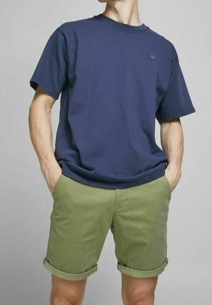 Mężczyzna w granatowym t-shircie z krótkim rękawem i oliwkowo-zielonych podwiniętych szortach, z rękami w kieszeniach, stoi na jednolitym tle.