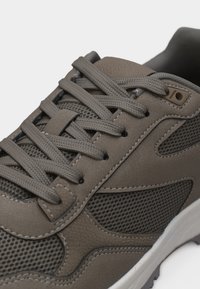 Baskets marron et grises avec panneaux en mesh, col rembourré et lacets gris sur fond blanc, montrant les détails latéraux et supérieurs.