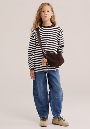 Jeune personne aux longs cheveux blonds portant un t-shirt rayé noir et blanc, un jean bleu troué, des baskets beiges et un sac à bandoulière marron avec une peluche.