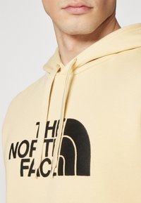 Svietlý žltý hoodie vyrobený z mäkkého materiálu. Obsahuje veľké čierne logo na prednej strane, šnúrky a klokanie vrecko.