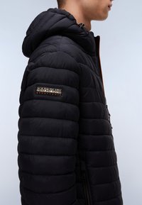 Black padded jacket with a hood, zip closure, side pockets, and a rectangular logo patch on the sleeve. Textured and lightweight material.