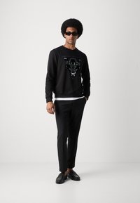 Sudadera negra con un gráfico de pantera texturizado, combinada con pantalones negros y zapatos negros. El conjunto se completa con gafas de sol oscuras.