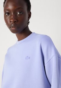 Lyse lilla genser laget av myk tekstil, med rund hals og en liten brodert logo på brystet. Jevn tekstur, avslappet passform.