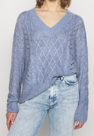 Light blue knitted sweater with a v-neck, featuring a diamond pattern and openwork details. Long sleeves with ribbed cuffs, paired with light jeans.