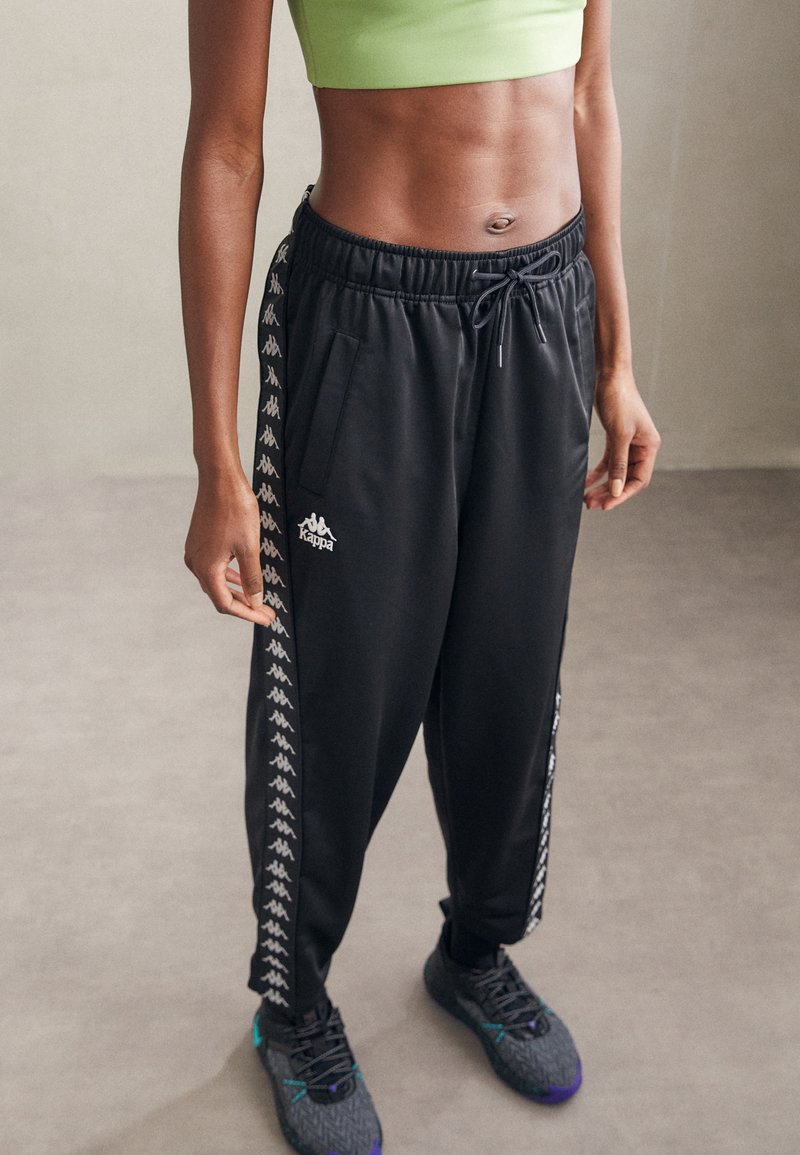 Pantaloni sportivi neri con vita elastica, caratterizzati dal logo Kappa e strisce bianche sui lati, abbinati a scarpe sportive nere dalla texture strutturata.