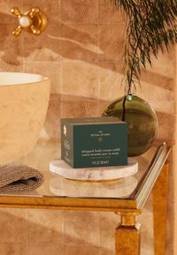 En refillboks med vispete kroppskrem i mørkegrønn med gulltekst, plassert på en marmorflate nær et gullbord, med en teksturert beige bakgrunn.