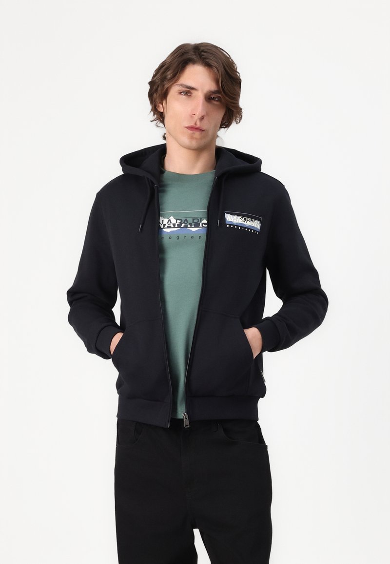 Czarna bluza z kapturem na zamek, z kieszenią kangurzą, posiadająca logo na klatce piersiowej i sznurkiem do regulacji kaptura, noszona na zielonej koszulce.