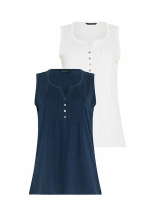 Frau mit lockigem Haar, die ein ärmelloses marineblaues Oberteil, marineblaue Shorts und weiße Turnschuhe trägt, vor einem hellen Hintergrund steht.