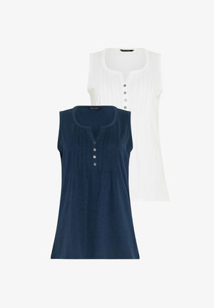 Vrouw met krullend haar, gekleed in een marineblauwe mouwloze top, marineblauwe korte broek en witte sneakers, staand tegen een lichte achtergrond.