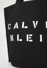 Juodas drobės rankinės maišas su didelėmis baltomis blokinėmis raidėmis, rašančiomis "CALVIN KLEIN." Su tvirtomis audinio rankenomis ir minimaliais siūlų akcentais.