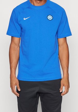 Mężczyzna noszący niebieską koszulkę Nike z logo klubu piłkarskiego Inter Mediolan, w połączeniu z czarnymi spodniami, stojący na jednolitym tle.