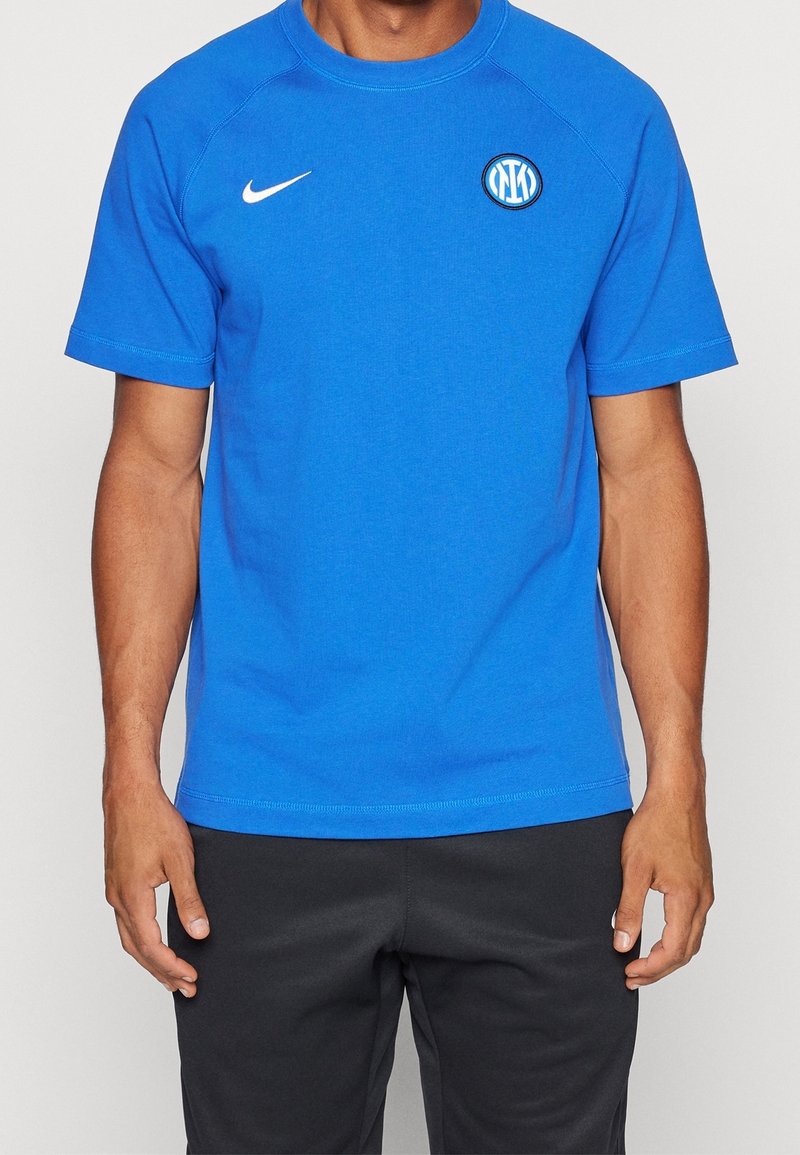 Homme portant un T-shirt bleu Nike avec le logo du club de football Inter Milan, associé à un pantalon noir, debout devant un fond uni.