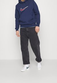 Man i marinblå hoodie med röd och vit logotyp, svarta uppvikta jeanstights och vita sneakers med mörkgrön logotyp som står framför en enhetlig bakgrund.