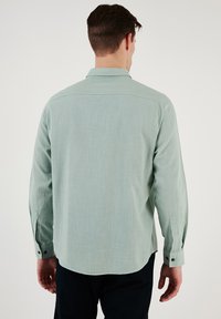 Lichtgroene blouse met lange mouwen, eenvoudige kraag, rugpasdetail en zichtbare knopenmanchetten. Gladde, gestructureerde stof.