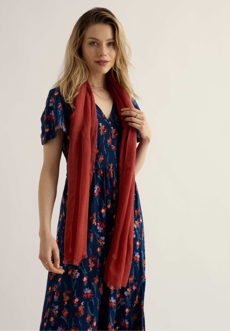 Femme aux cheveux blonds portant une robe marine à motifs floraux et une longue écharpe rouge, tenant légèrement l'écharpe près de son épaule.