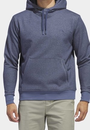 Man draagt een blauwe hoodie met een voorzak en een logo op de borst, gecombineerd met beige broek, staand met ontspannen armen.