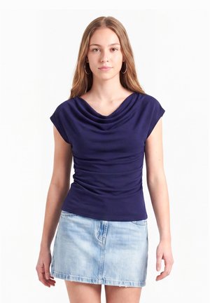 Jonge vrouw met lang lichtbruin haar, gekleed in een marineblauw gedrapeerd topje en een lichte denim minirok, staand tegen een witte achtergrond.