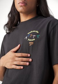 Osoba v černém tričku s barevnými figurkami a textem "Mládež je budoucnost" a "PUMA," ruka položená na hrudi.