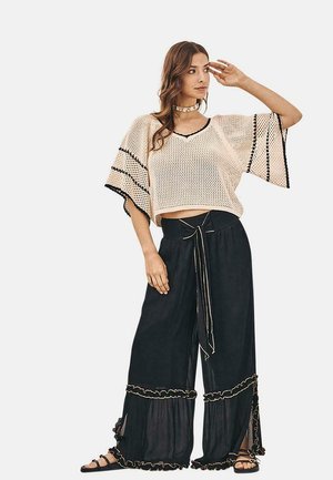 Top de punto beige con ribete negro; corte holgado. Pantalones negros de pierna ancha con dobladillo con volantes y detalle de cintura con lazo. Mujer posando con la mano en el cabello.