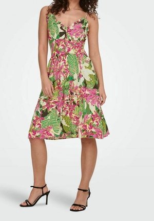 Frau trägt ärmelloses, knielanges Kleid mit grünem, pinkem und weißem tropischem Blumenmuster und schwarze, hochhackige Sandalen mit Riemchen.