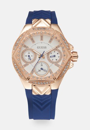 Montre en or rose avec un bracelet en silicone bleu texturé, boîtier octogonal, accents de cristal et un cadran blanc avec trois sous-cadrans.