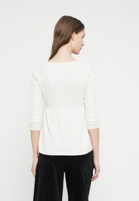 Λευκό πλεκτό πουλόβερ με μανίκια τριών τετάρτων, με υφασμένο σχέδιο καλωδίου και φαρδύ στη μέση. Λείο φινίρισμα, χαλαρή εφαρμογή.