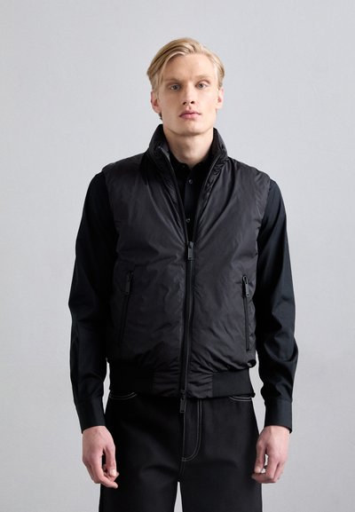Gilet imbottito nero con colletto alto, zip frontale e due tasche laterali. Indossato sopra una maglietta nera, presenta un design elegante e moderno.