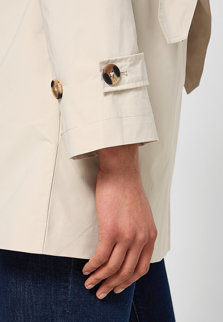Chaqueta impermeable beige con un acabado ligeramente brillante, que presenta grandes botones marrones y negros en los puños, y un corte relajado y entallado.