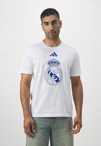 Hvid bomulds t-shirt med et blåt Real Madrid-logo og krone-design på fronten. Korte ærmer og rund hals.