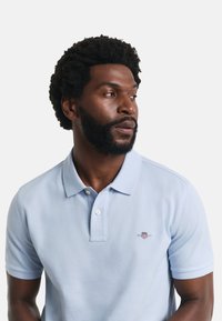 Jasnoniebieska koszulka polo z klasycznym kołnierzykiem, krótkimi rękawami i haftowanym logo na lewej piersi. Gładka, teksturowana bawełniana tkanina.