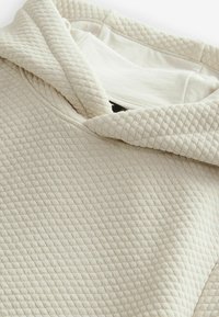 Beżowy hoodie o fakturowanym, pikowanym wzorze. Kaptur jest lekko za duży, z gładką podszewką, z delikatnymi detalami przeszycia.