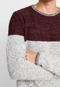 Pull tricoté bordeaux et gris avec un design en blocs de couleurs, présentant une finition texturée et un col et des poignets côtelés.