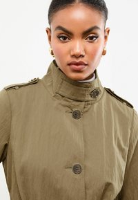 Vrouw in een hooggesloten olijfgroene jas met knopen, gouden hoepeloorbellen en een neutrale make-uplook, die recht naar voren kijkt tegen een witte achtergrond.