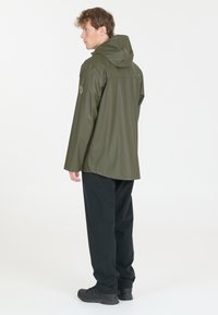 Olivgrüne, wasserdichte Jacke mit Kapuze, die eine glatte Textur und einen geraden Schnitt aufweist, getragen über einer schwarzen Hose und stabilen schwarzen Schuhen.