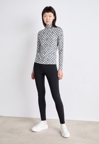 Langärmliges, Rollkragenoberteil in Weiß mit einem schwarzen geometrischen Muster, kombiniert mit schwarzen Leggings und weißen Turnschuhen, vor einem neutralen Hintergrund.