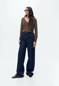 Maglione a maniche lunghe marrone, jeans ampi a vita alta blu scuro, e scarpe nere basse con una leggera lucentezza. Gli occhiali da sole incorniciano il viso.
