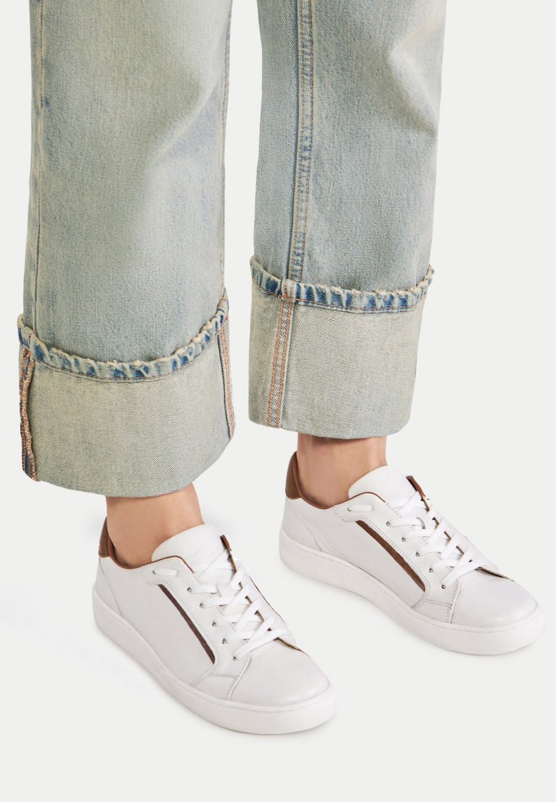 Valged nahast tennisetossud pruuni aktsendiga, madala lõikega disain, tasase tallaga ja esiosas seotud nööpidega, koos helesiniste lühikeste teksadega.