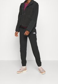 Svart fleecejacka med tryckknappar och två framfickor, kombinerad med svarta sweatpants med en logotyp. Ljusfärgade sneakers med snörning.