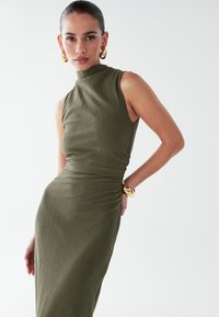 Robe côtelée vert olive avec col montant et design sans manches, présentant des fronces à la taille et une texture lisse. Accent bracelet doré.