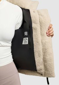 Beżowa bluza polarowa z czarnym wnętrzem. Posiada metkę z napisem "Ciepły uścisk wykonany z tkaniny", podkreślającą miękką teksturę i warstwowy design.