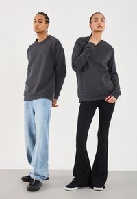 To personer står side om side iført mørkegrå hettegensere; den ene i lys blå jeans og svarte sneakers, den andre i svarte flagrende bukser og svarte sko.