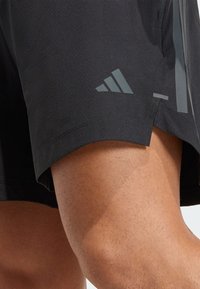 Pantalones cortos deportivos negros con un acabado texturizado, con un diseño reflectante de tres rayas y un logo de adidas en gris oscuro en el dobladillo.