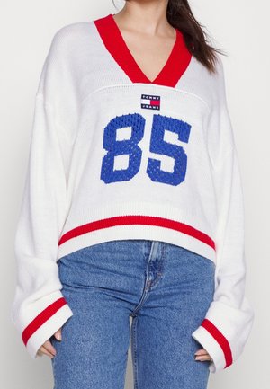 Pull en maille blanche avec col en V rouge et poignets rayés, arborant une broderie bleue "85" et un patch logo TOMMY JEANS sur le devant.