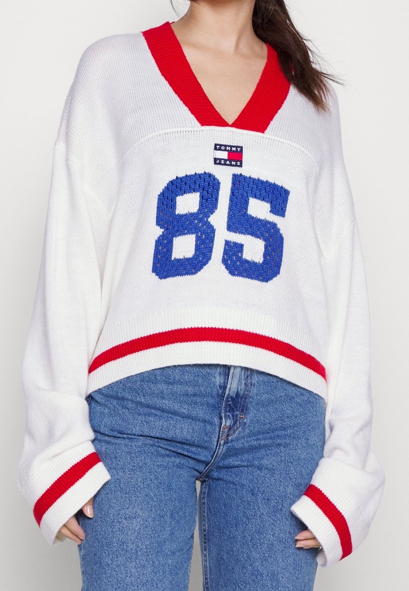 Bílý pletený svetr s červeným výstřihem do V a pruhovanými manžetami, s modrou výšivkou "85" a patchem loga TOMMY JEANS na přední straně.