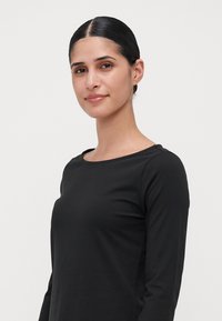Young woman with dark hair tied back wearing a black long-sleeve shirt, standing against a plain white background.