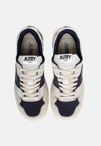 Baskets blanches et navy présentant un mélange de matériaux en cuir et en daim, avec des accents perforés et une semelle en caoutchouc. Lacets et logo visibles.