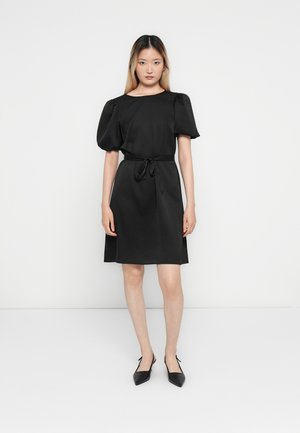 Femme debout portant une robe noire longueur genou avec des manches courtes bouffantes, une ceinture nouée à la taille et des chaussures à talons aiguille noires avec un talon ouvert.
