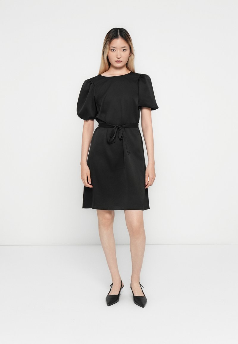 Femme debout portant une robe noire longueur genou avec des manches courtes bouffantes, une ceinture nouée à la taille et des chaussures à talons aiguille noires avec un talon ouvert.